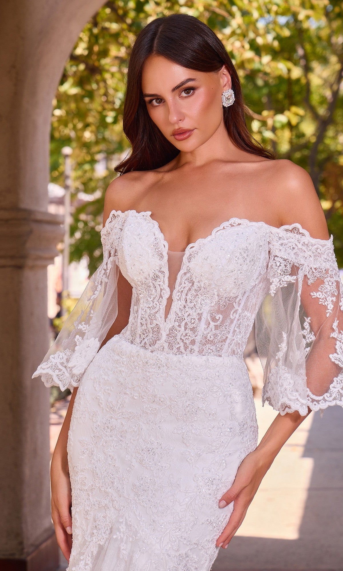 A woman wearing the Ladivine CD659W long wedding dress, featuring an off-the-shoulder lace design with sheer sleeves and an elegant mermaid skirt, stands outdoors by an archway surrounded by greenery.